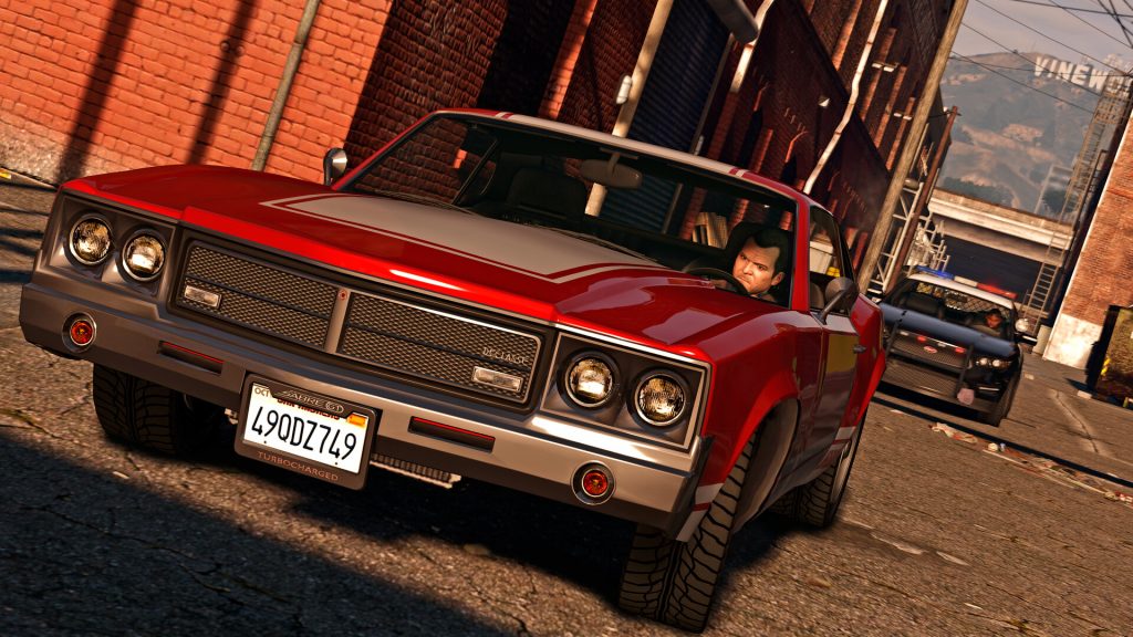 A red muscle car speeding through a sunlit alley with a police cruiser close behind